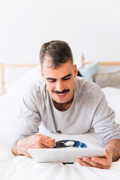 Smiling man eating yogurt and watching film on tablet