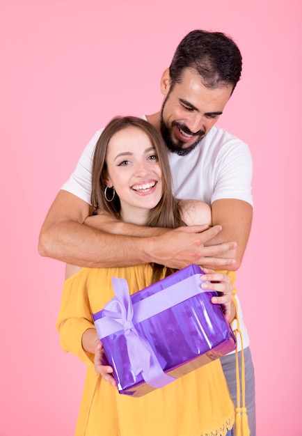 Smiling man embracing her girlfriend holding present against pink backdrop