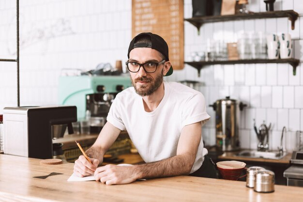 鉛筆とメモ帳でバーカウンターの後ろに立って、カフェで夢のようにカメラで見ている眼鏡とキャップの笑顔の男