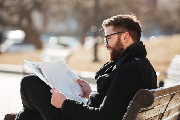 Улыбающийся человек в очках читает газету на скамейке на открытом воздухе