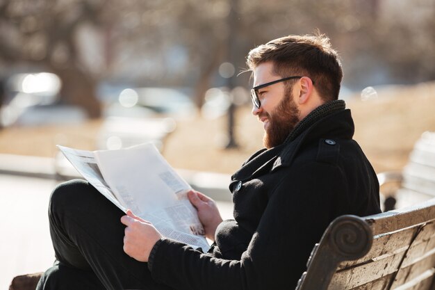 Smiling man in glasses reading newspaper on the bench outdoors