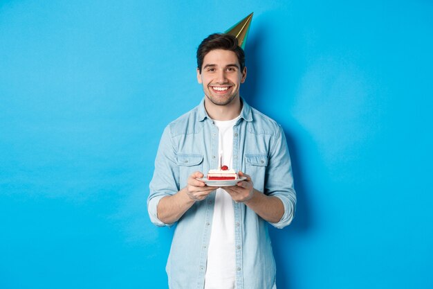 Smiling man holding b-day cake and wearing birthday party hat, celebrating over blue background.