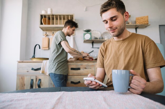 彼の友人が台所のカウンターで食べ物を準備しながら携帯電話とコーヒーカップを保持している笑みを浮かべて男