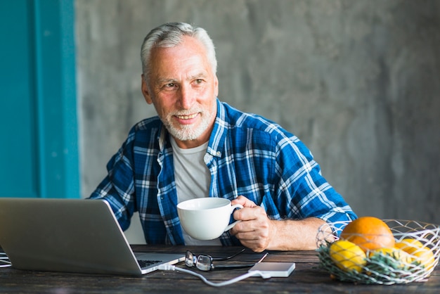 Smiling man holding coffee cup looking away with laptop on table