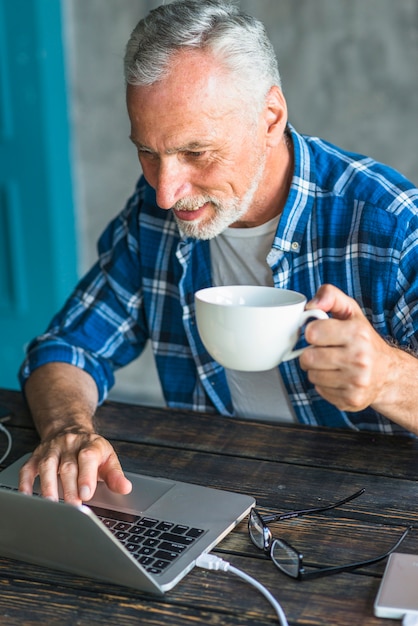 Smiling man holding coffee cup using laptop over the wooden desk