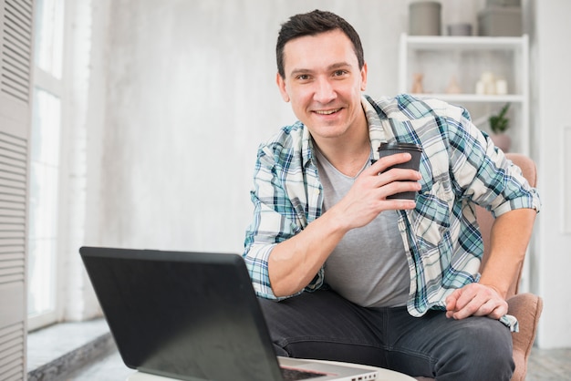 Улыбающийся человек, держащий чашку напитка на стуле возле ноутбука дома