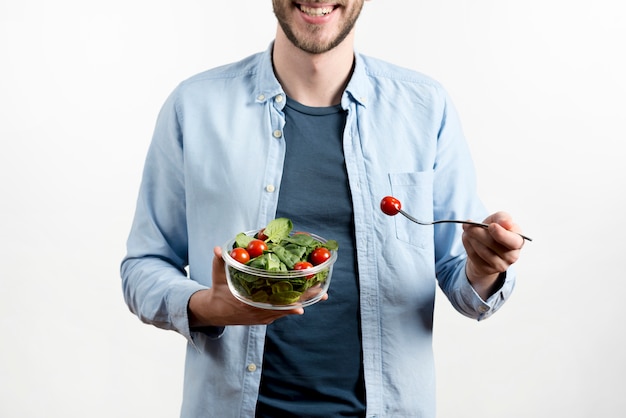 Smiling man holding fork with cherry tomato and bowl of salad against white background