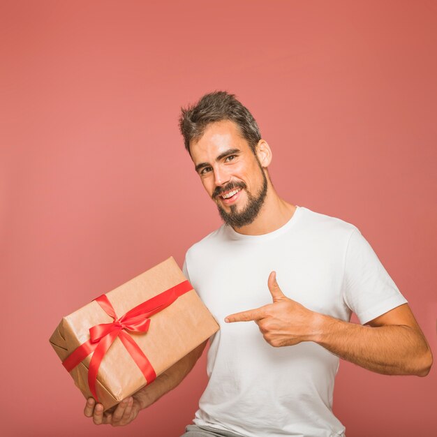 Smiling man holding gift box pointing finger against colored backdrop