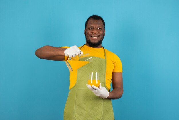 Smiling man holding glass while pouring fruit juice from carafe , stands over blue wall. 