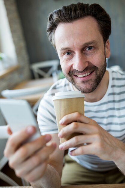 Smiling man holding mobile phone and coffee cup in coffee shop