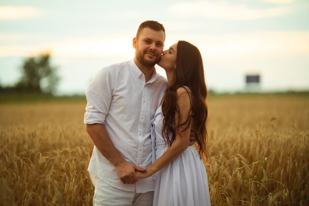Smiling man hugging her pretty wife while standing behind her on a wheat field during evening sunset. Love concept