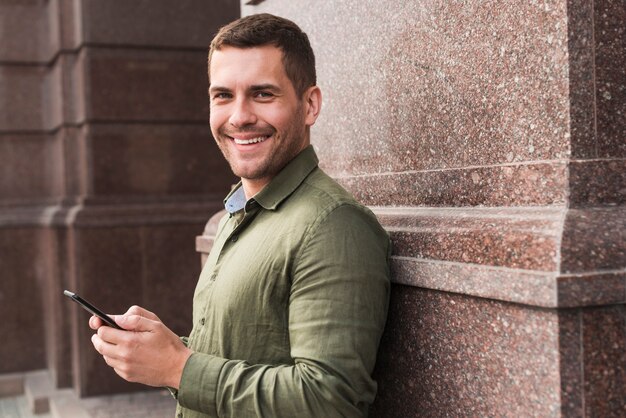 Smiling man leaning on wall holding cellphone and looking at camera