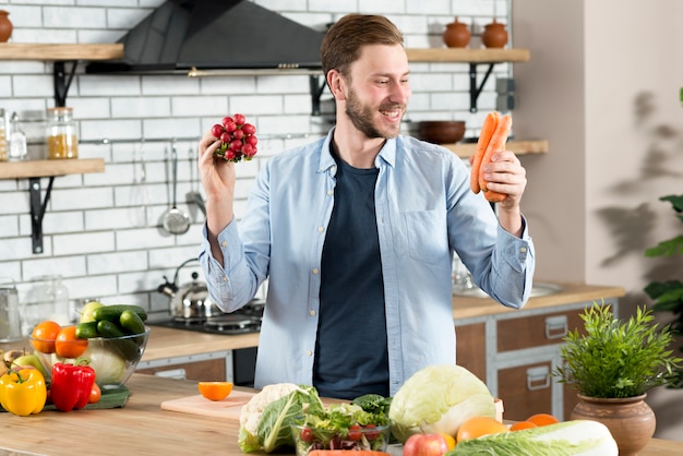 Smiling man looking at orange carrot standing behind kitchen counter