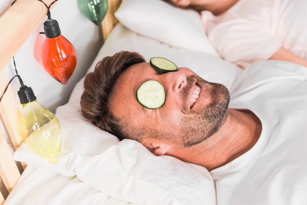 Smiling man lying on bed with cucumber slice over eyes