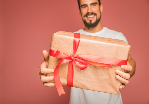 Smiling man offering gift box with red ribbon bow against colored backdrop