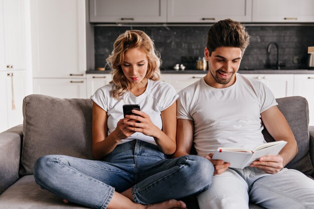 Smiling man reading book in living room. Indoor portrait of curly young woman holding smartphone.