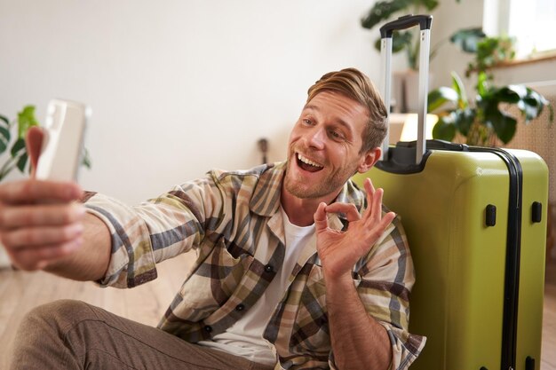 Smiling man shows okay sign poses near luggage goes on holiday takes selfie on mobile phone