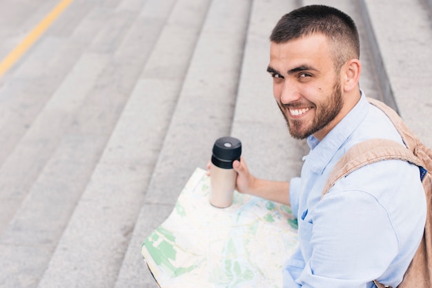 Foto gratuita uomo sorridente che si siede sulla mappa della tenuta della scala e sulla tazza eliminabile che esaminano macchina fotografica