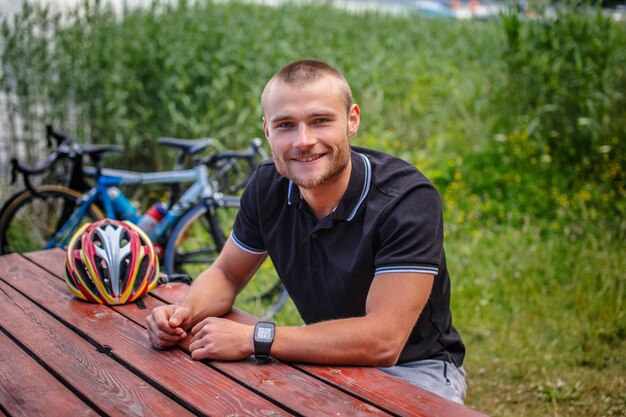 Smiling man sitting at table on lake shore with sport bycicles on background.