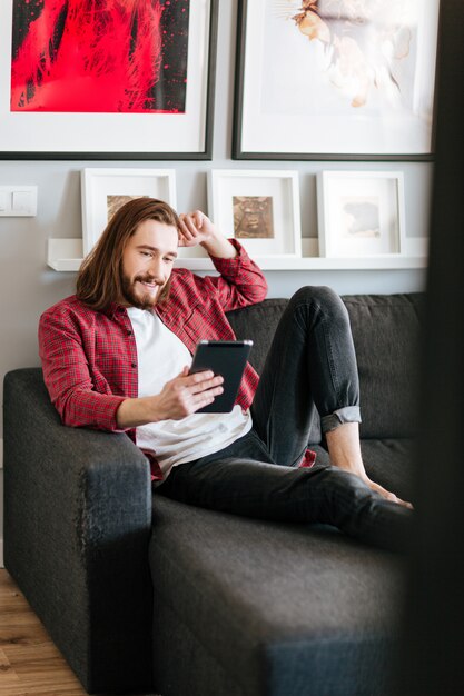 笑みを浮かべて座っている男と自宅のソファの上のタブレットを使用して