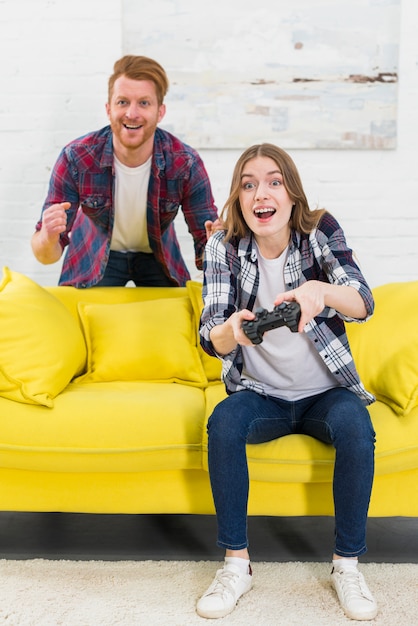 Улыбающийся человек, стоящий за возбужденной женщиной, играющей в видеоигру в гостиной