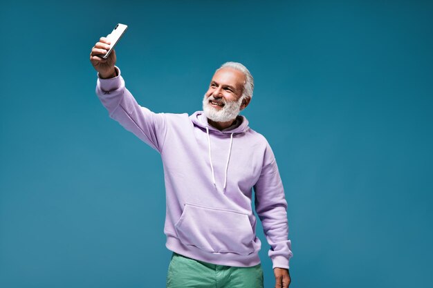 Smiling man in stylish outfit takes selfie on blue background Cool guy in modern hoodie and green plants smiling on isolated backdrop