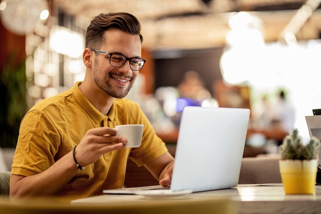 Улыбающийся мужчина бродит по сети на ноутбуке, попивая кофе в баре