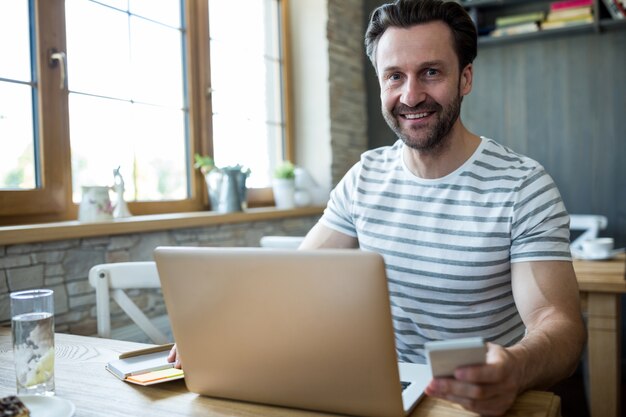 Smiling man using his laptop and mobile phone in coffee shop