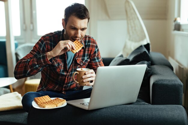 Smiling man using laptop while drinking coffee and eating waffle in the living room