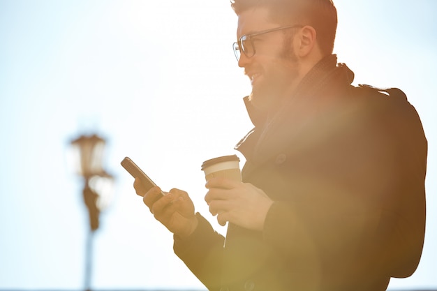 Улыбающийся человек с помощью мобильного телефона и пить кофе на открытом воздухе