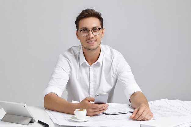 Smiling man wears white formal shirt and round spectacles, holds mobile phone, messages, drinks coffee, writes sketches, has positive expression. Well educated designer uses modern technologies