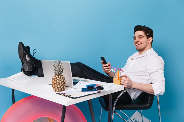 smiling man in white shirt and pants is sitting with his legs on table against blue space. brunette guy holding phone and cocktail. employee posing with laptop, pineapple and inflatable circle.
