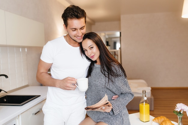 Smiling man with cup of coffee embracing brunette woman holding newspaper