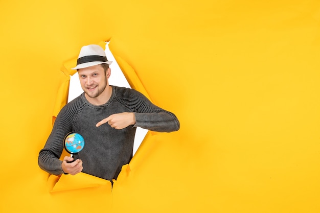 Free photo smiling man with a hat holding small globe and pointing a small globe in a torn on yellow wall