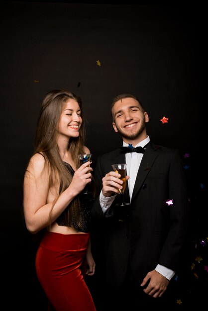 Smiling man and woman in dinner jacket and evening wear with glasses of drinks near confetti 