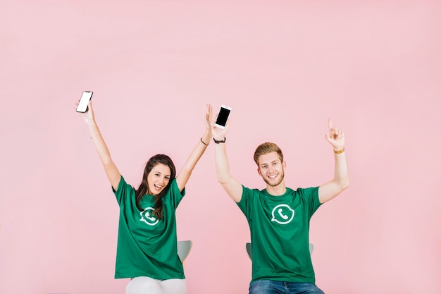 Free photo smiling man and woman with mobile phone raising their arms over pink background