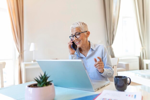 Smiling mature beautiful business woman with white hair working on laptop in bright modern home office