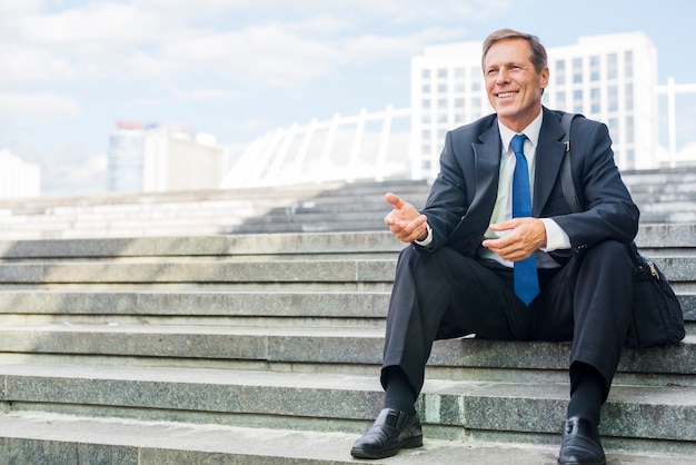 Smiling mature businessman making hand gesture sitting on staircase