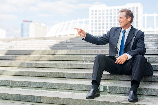 Free photo smiling mature businessman pointing at something while sitting on staircase