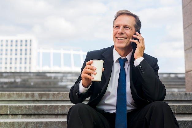 携帯電話で話すコーヒーのカップで笑顔の成熟したビジネスマン