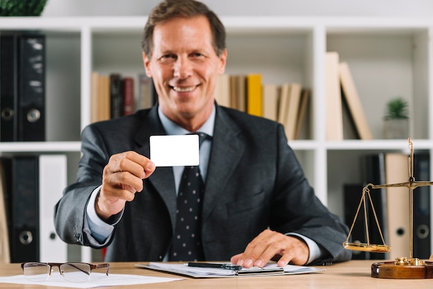 Smiling mature lawyer showing blank visiting card sitting in the court room