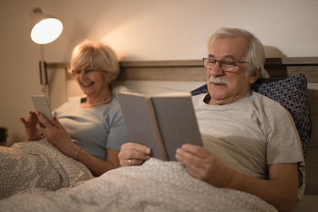 Smiling mature man lying down in bed and reading a book while his wife is using mobile phone