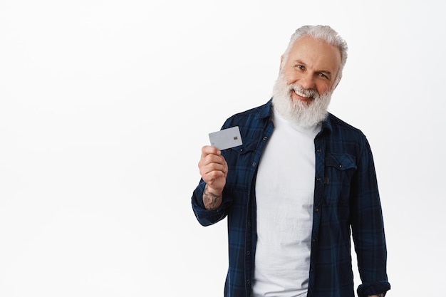 Smiling mature man with tattoos shows credit card, laughs and looks happy at camera, recommends bank, paying contactless, order with card, stands over white background