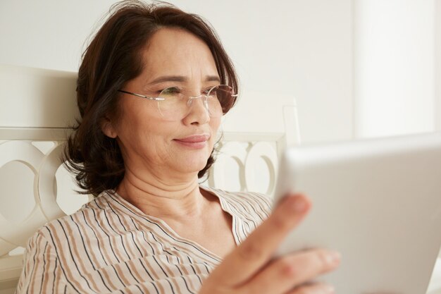 Smiling mature woman in glasses using digital tablet pc lying on her bed in a bedroom