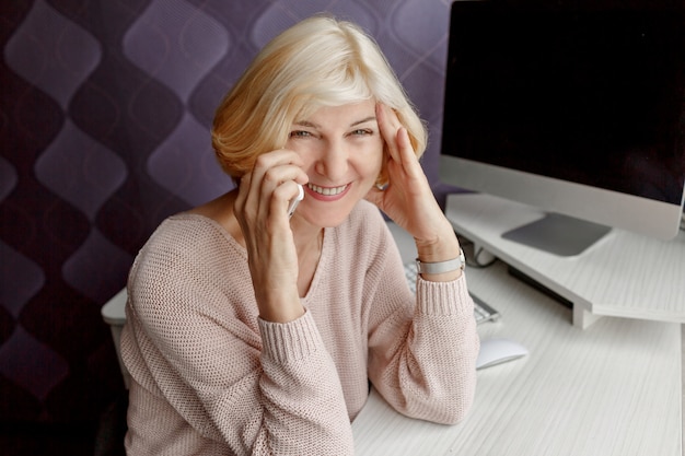 Smiling mature woman using mobile phone while working by computer at home