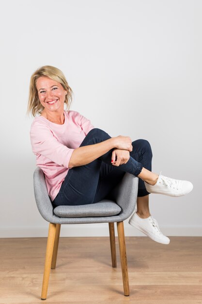Smiling mature woman in white canvas shoes sitting on gray chair against white wall