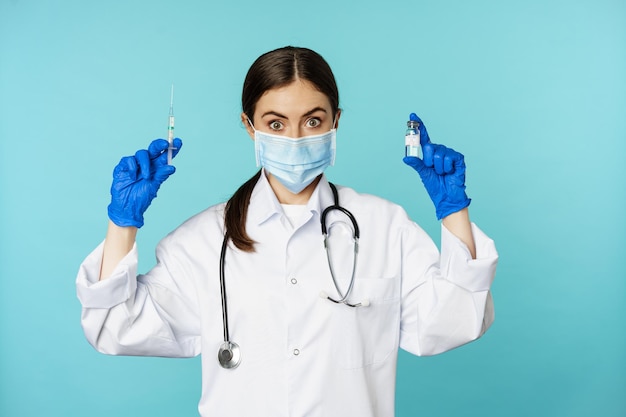 Улыбающийся врач медицинского персонала в маске и резиновых перчатках показывает шприц и вакцину от covid p...
