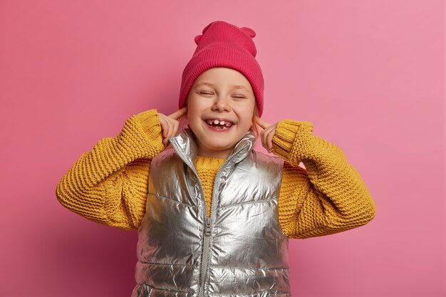 Smiling merry little child plugs ears with fore fingers, doesnt want to hear noisy neighbours, wears hat, knitted sweater and vest, avoids loud sound, smiles broadly, shows teeth isolated on pink wall