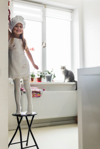 Smiling messy little girl in chef hat and apron standing on table at home