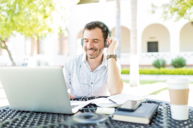 Maschio adulto metà sorridente che usa le cuffie collegate al computer portatile mentre fa il telelavoro in giardino